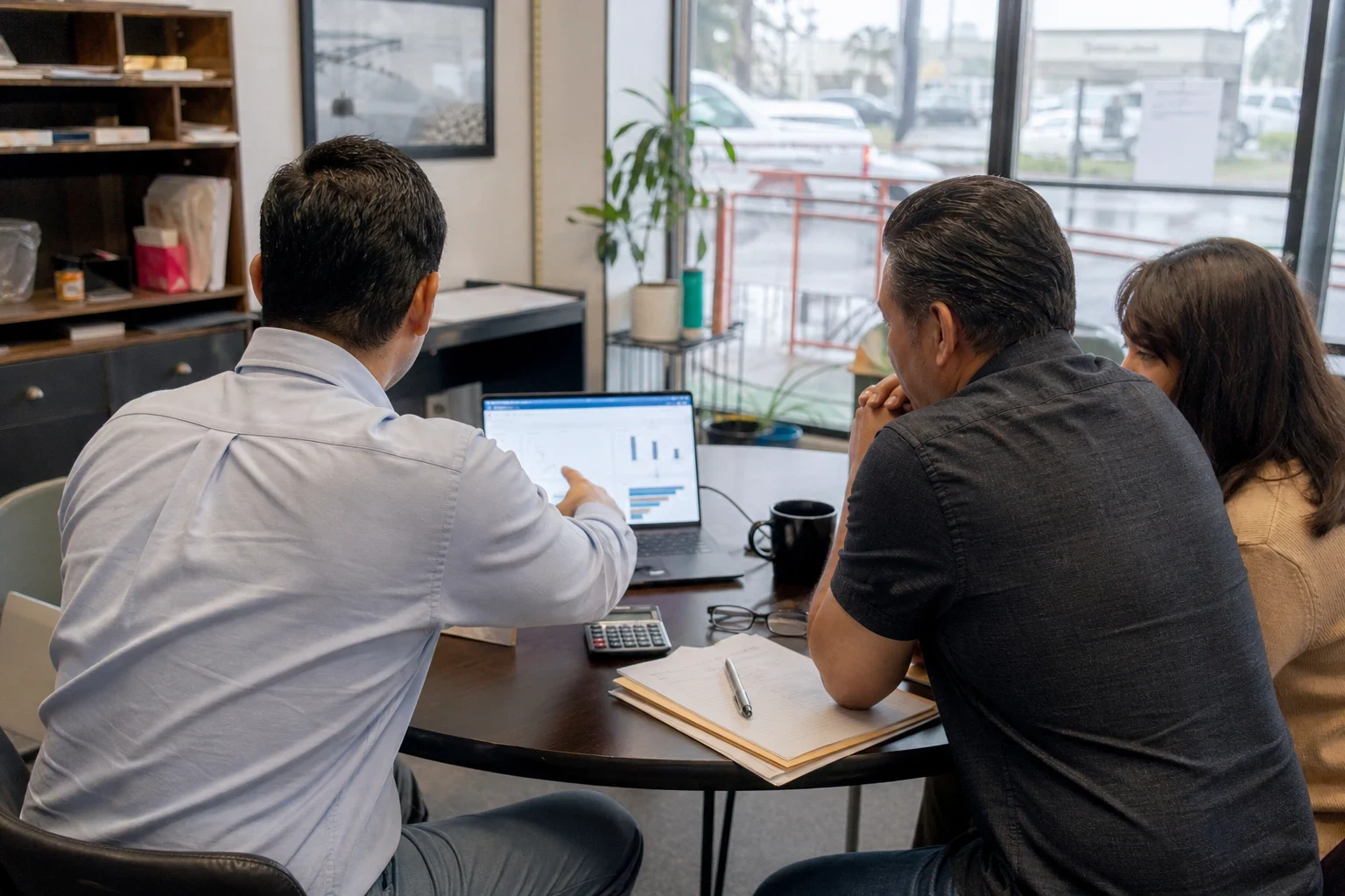 Agent in a light blue button-down points to charts on a laptop while a man and woman sit beside him at a round table in a storefront office with an overcast street view